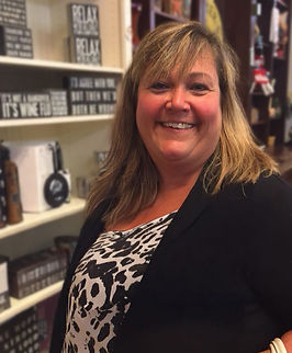 A smiling woman with shoulder-length hair stands in a store filled with decorative signs and products, promoting a warm, welcoming atmosphere.