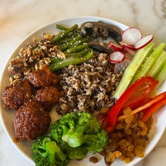 A colorful plate featuring three falafel-style balls, broccoli, wild rice, asparagus, mushrooms, radishes, celery, red bell pepper, walnuts, and raisins.