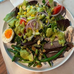Fresh salad featuring mixed greens, cherry tomatoes, green beans, corn, red onions, avocado dressing, and sliced boiled egg, presented on a white plate.