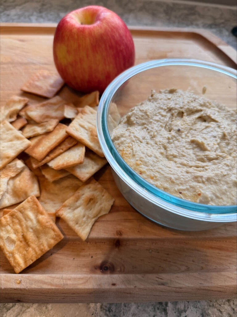 Fresh apple, hummus in a glass bowl, and crispy crackers arranged on a wooden cutting board, showcasing a healthy snack option.